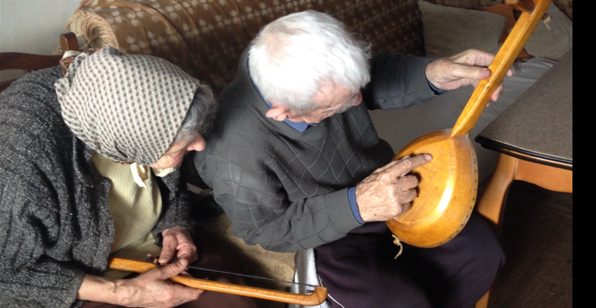 The Šolajas with their family gusle. Nadanići, Bosnia, 2012. Courtesy of Safet HadžiMuhamedović. An elderly woman holds a curved stick with strings (the bow of an instrument). Next to her a white-haired man points to the wooden back of a long-necked musical instrument.