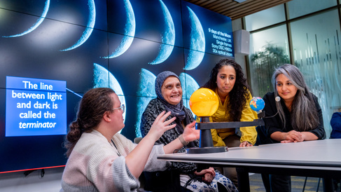 Three women of different ages sit at table with sun, earth and moon model. Emma crouches by the model explaining.