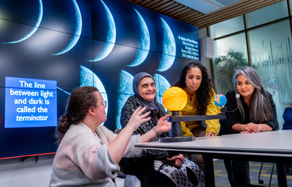 Three women of different ages sit at table with sun, earth and moon model. Emma crouches by the model explaining.