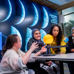 Three women of different ages sit at table with sun, earth and moon model. Emma crouches by the model explaining.