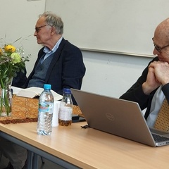 Ochs (seated right) smiles, chin-on-hands, looking towards companion SR facilitators. Head pointed away from the camera, his kippah is visible. Demiri sits at the left, mouth open in speech. Between is Ford, books splayed open.