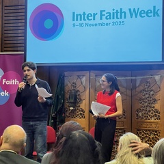 Two young people holding microphones stand in front of a projected Inter Faith Week logo, audience facing them.