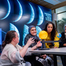 Three women of different ages sit at table with sun, earth and moon model. Emma crouches by the model explaining.