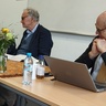 Ochs (seated right) smiles, chin-on-hands, looking towards companion SR facilitators. Head pointed away from the camera, his kippah is visible. Demiri sits at the left, mouth open in speech. Between is Ford, books splayed open.