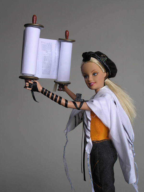 Blonde plastic doll with prayer shawl and bindings, holding torah scroll
