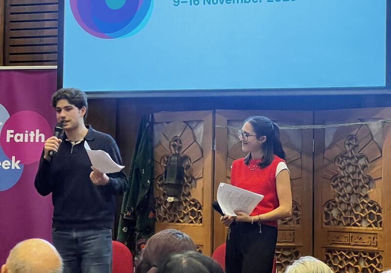 Two young people holding microphones stand in front of a projected Inter Faith Week logo, audience facing them.