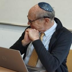 Ochs (seated right) smiles, chin-on-hands, looking towards companion SR facilitators. Head pointed away from the camera, his kippah is visible. Demiri sits at the left, mouth open in speech. Between is Ford, books splayed open.