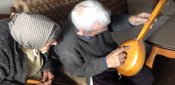 The Šolajas with their family gusle. Nadanići, Bosnia, 2012. Courtesy of Safet HadžiMuhamedović. An elderly woman holds a curved stick with strings (the bow of an instrument). Next to her a white-haired man points to the wooden back of a long-necked musical instrument.