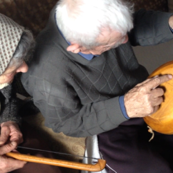 An elderly woman holds a curved stick with strings (the bow of an instrument). Next to her a white-haired man points to the wooden back of a long-necked musical instrument.