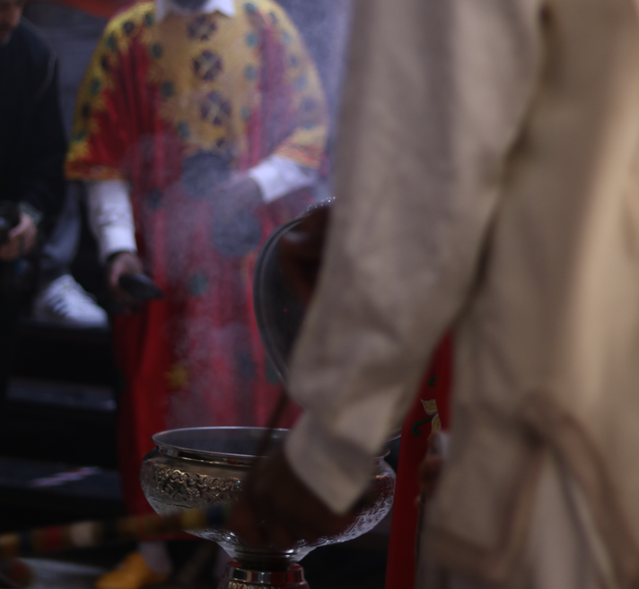 In the foreground, we glimpse drumstick and drum. Opposite the unseen player a man in ceremonial attire holds a percussive object. Between them a metal bowl gleams. Is it from here the smoke of incense rises?